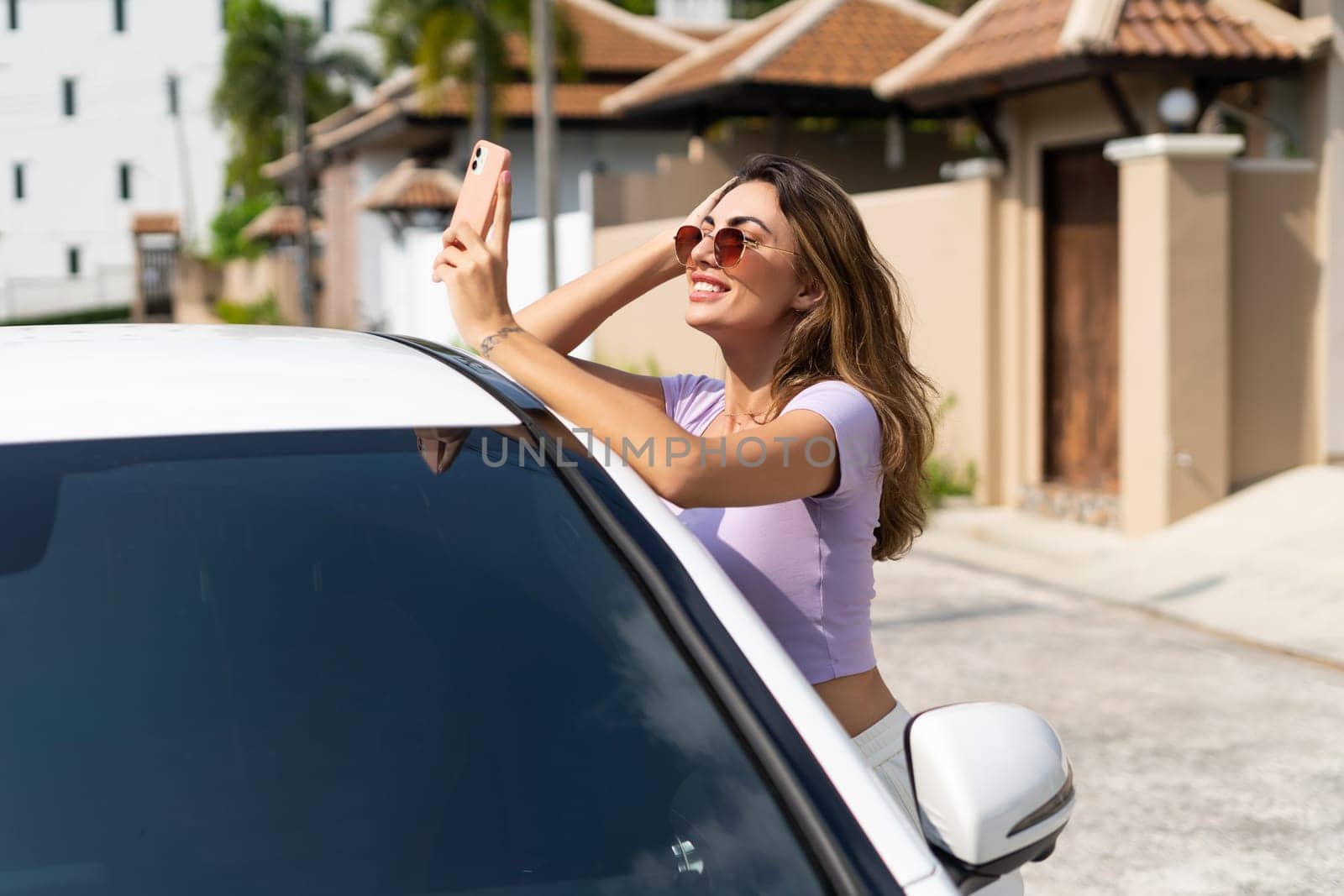 Successful smiling fit tanned attractive woman in casual wear is using her smart phone while standing near luxury modern car outdoors.