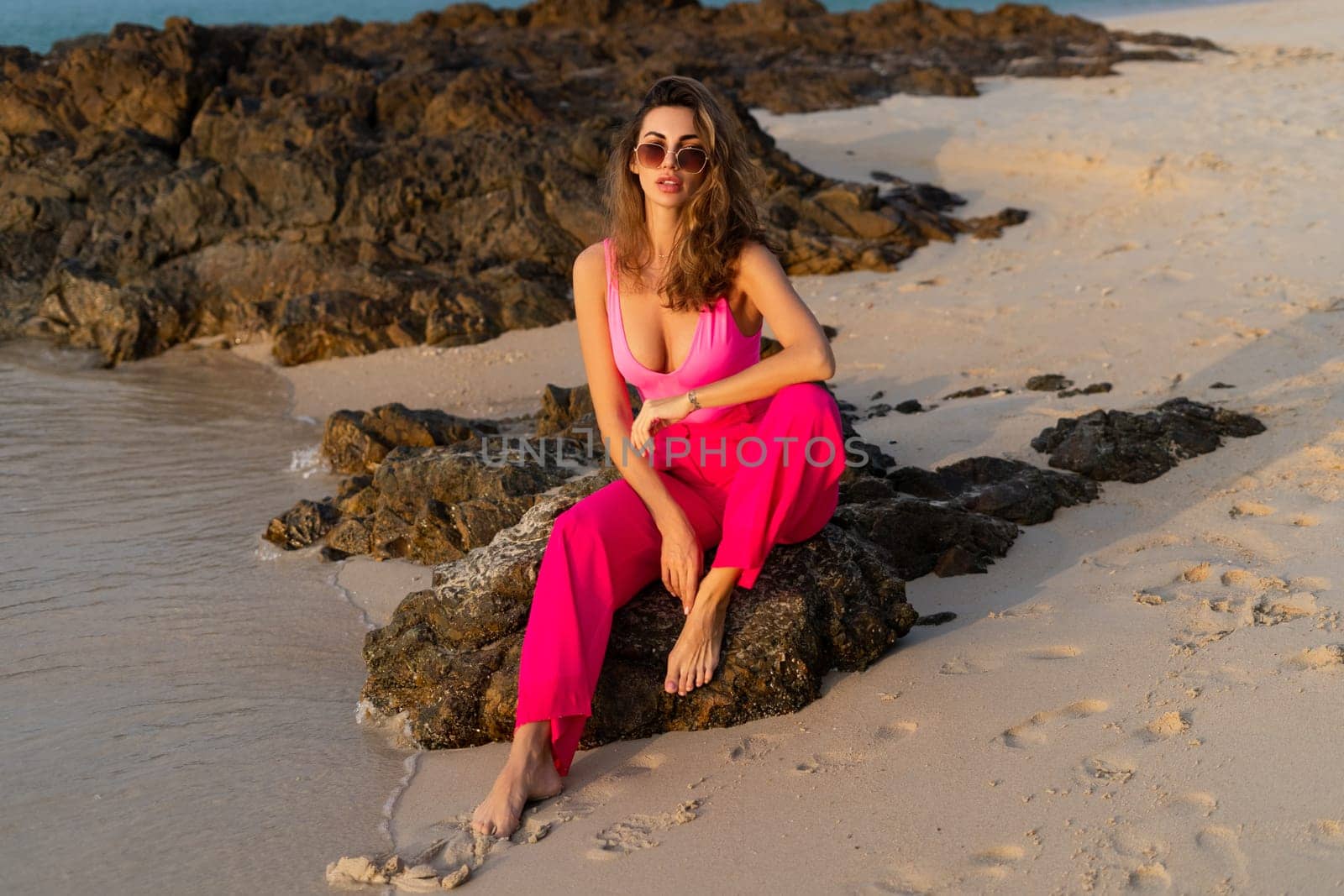 Fashion stylish beautiful sensual young woman, slim, in trendy bright pink pants and bodysuit on a tropical beach at sunset posing in sunglasses