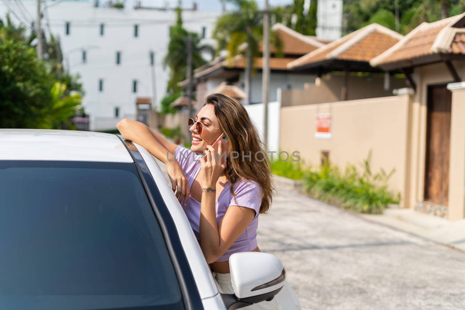 Successful smiling fit tanned attractive woman in casual wear is using her smart phone while standing near luxury modern car outdoors.