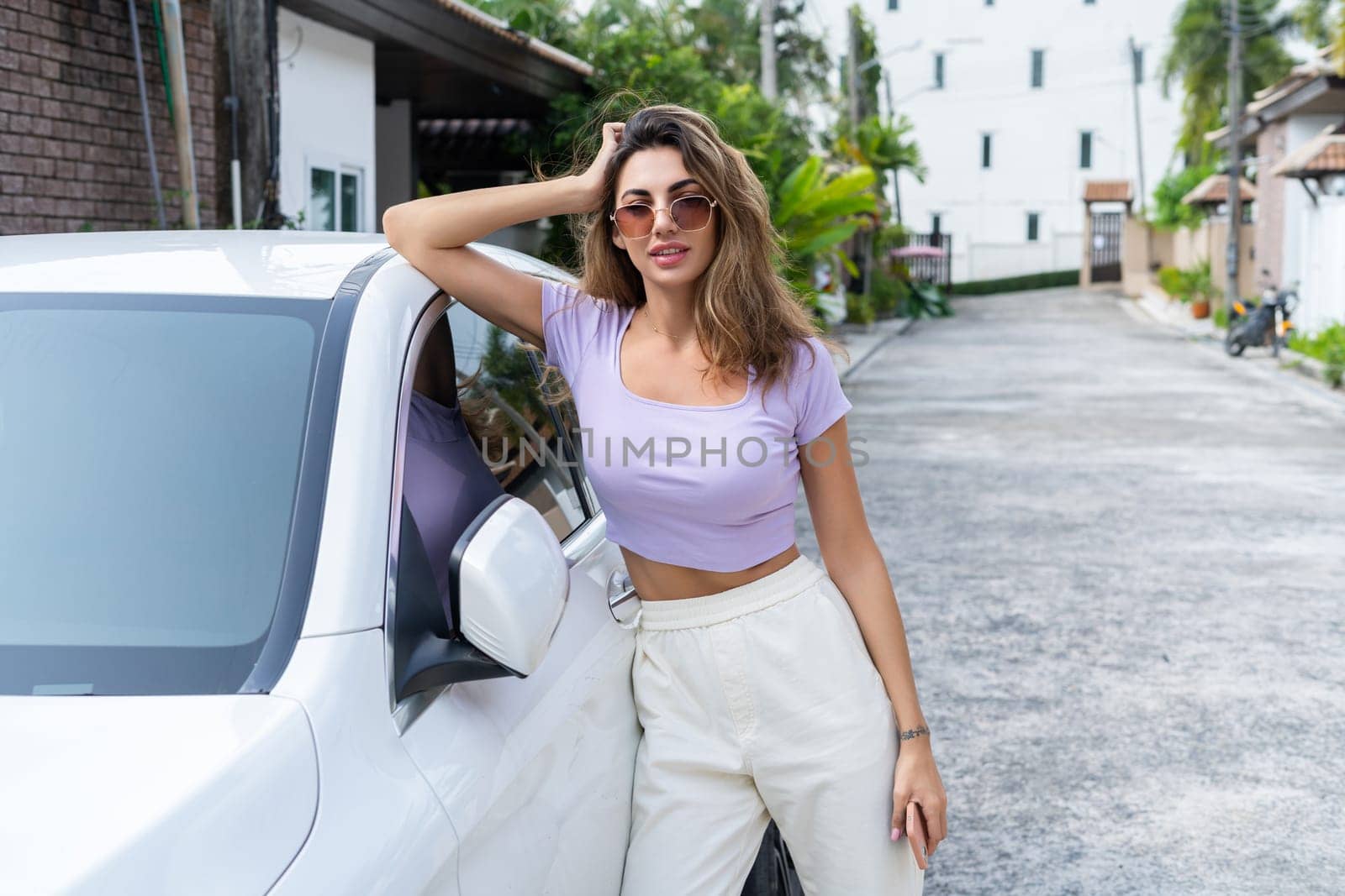 Successful smiling fit tanned attractive woman in casual wear is using her smart phone while standing near luxury modern car outdoors.