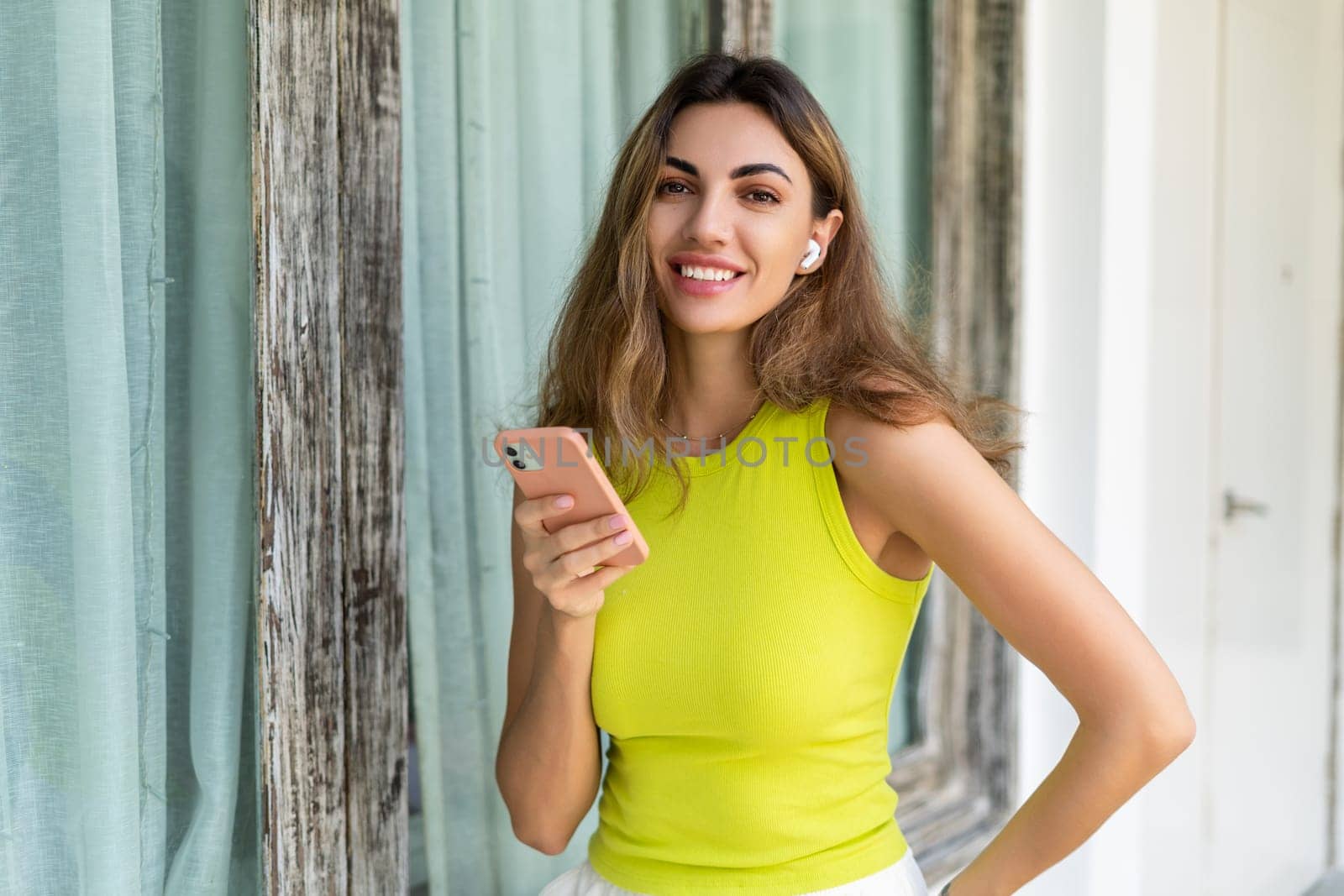 Welcome. Portrait of cheerful woman standing at modern villa with smartphone and headphones holding phone with happy thoughtful smile