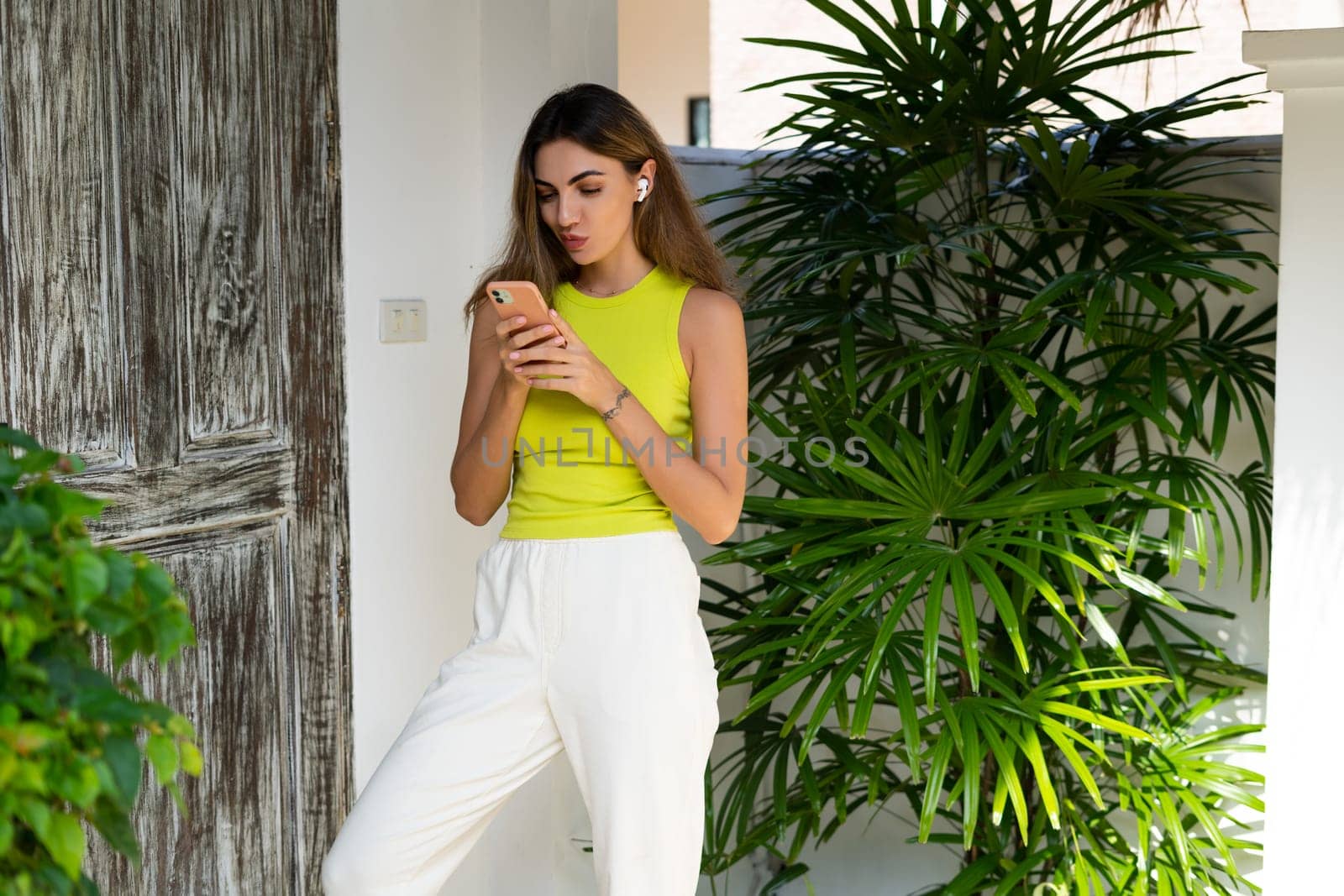 Welcome. Portrait of cheerful woman standing in doorway of modern villa with smartphone and headphones looking at screen with thoughtful smile