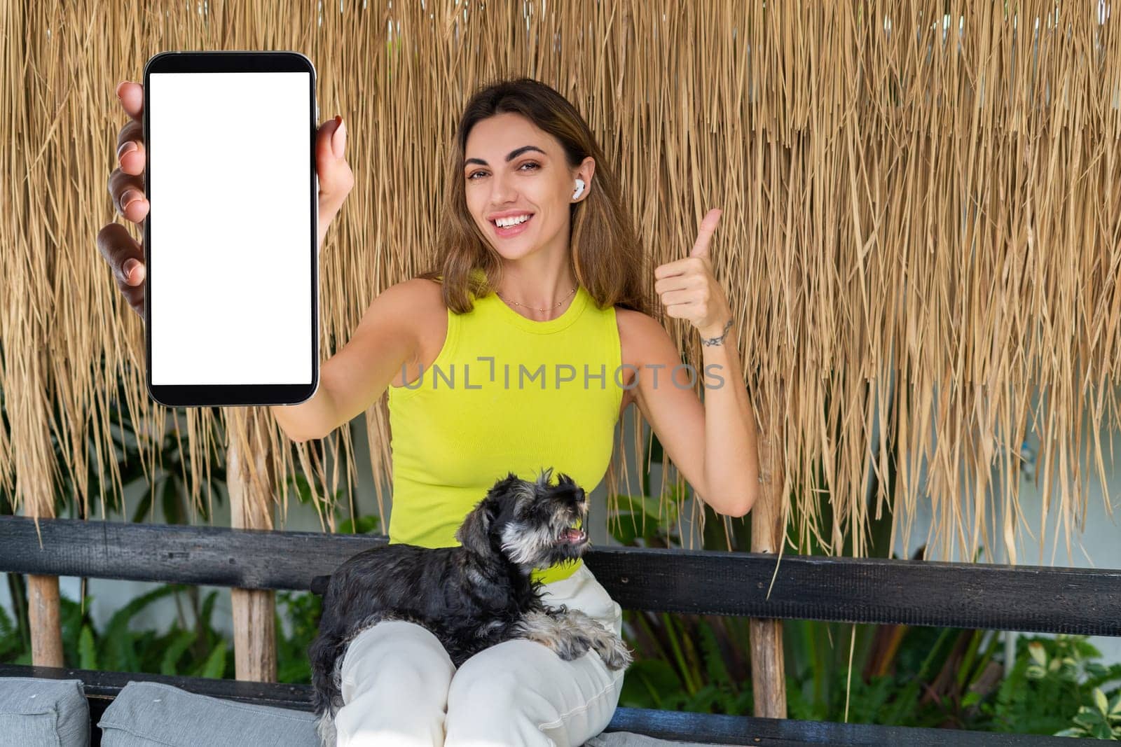 Portrait of cheerful woman standing outdoor with smartphone and headphones, with miniature schnauzer puppy dog, holding phone with white empty big screen with happy smile by kroshka_nastya