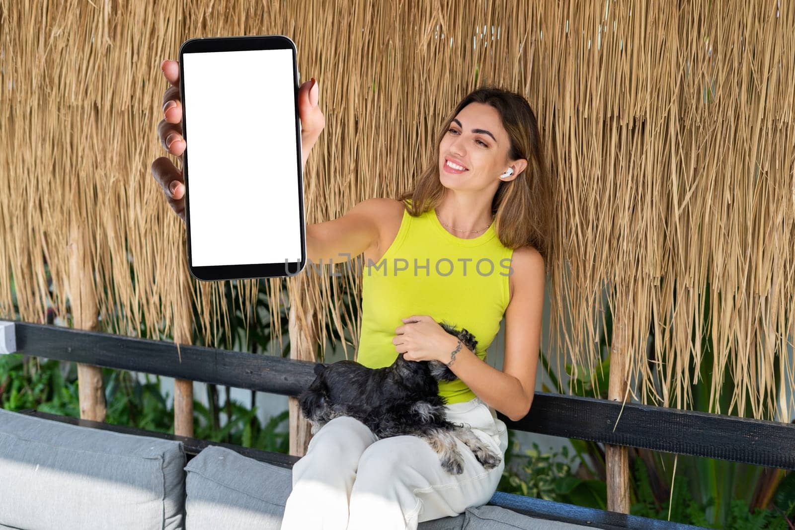 Portrait of cheerful woman standing outdoor with smartphone and headphones, with miniature schnauzer puppy dog, holding phone with white empty big screen with happy smile