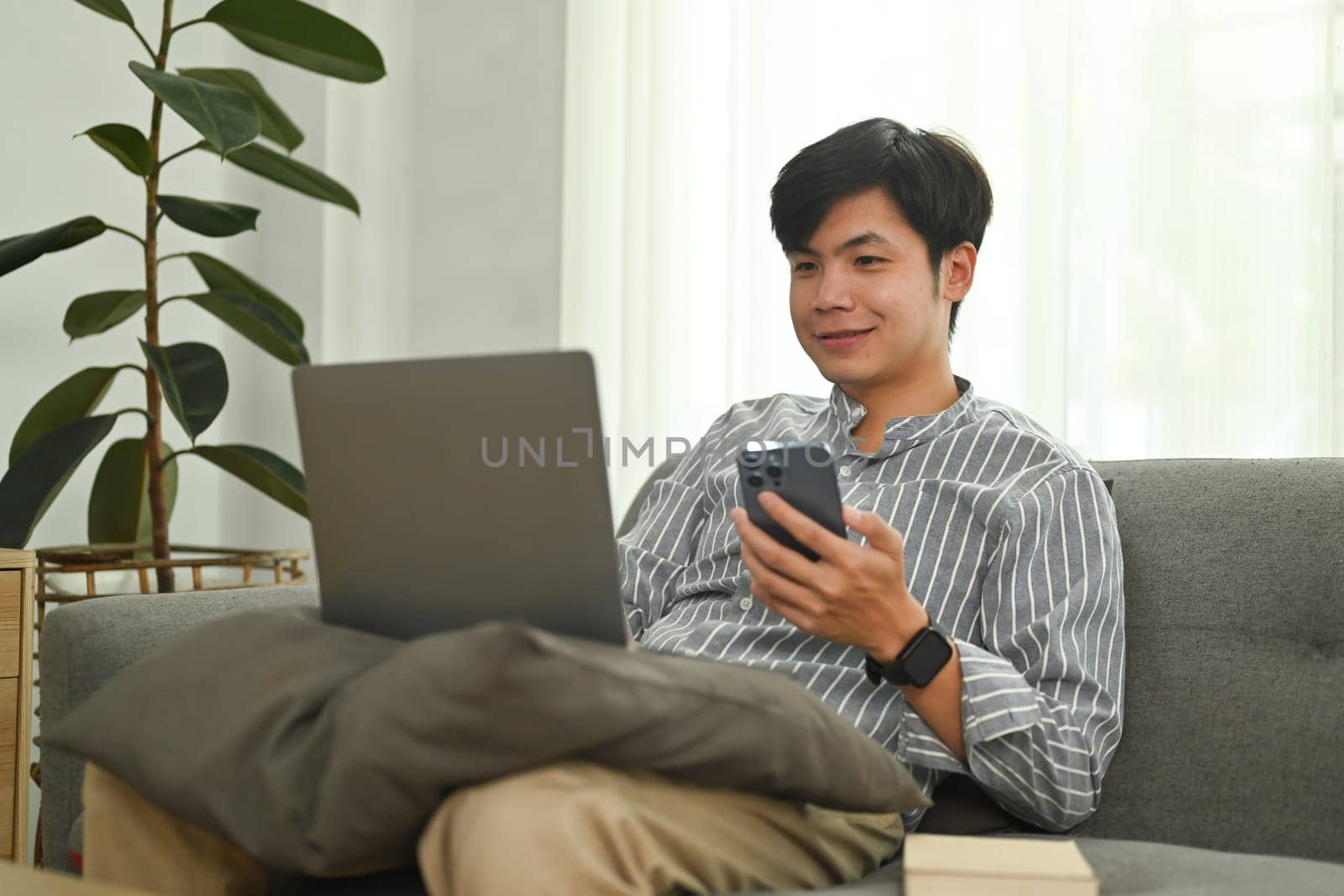 Cheerful asian man hand holding mobile phone and looking at laptop screen. People, technology and lifestyle concept.