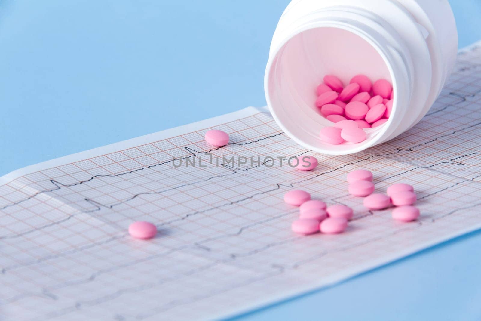 A large handful of pink pills poured out of a white jar on an electrocardiogram of the heart, on a blue background. The concept of a healthy lifestyle and timely medical examination