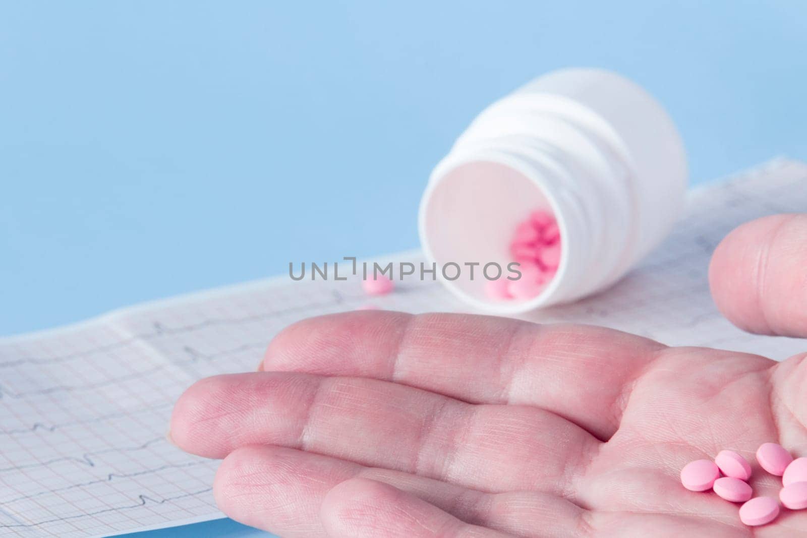 A hand pours a handful of pink pills from a white jar onto an electrocardiogram of the heart, on a blue background. The concept of a healthy lifestyle and timely medical examination