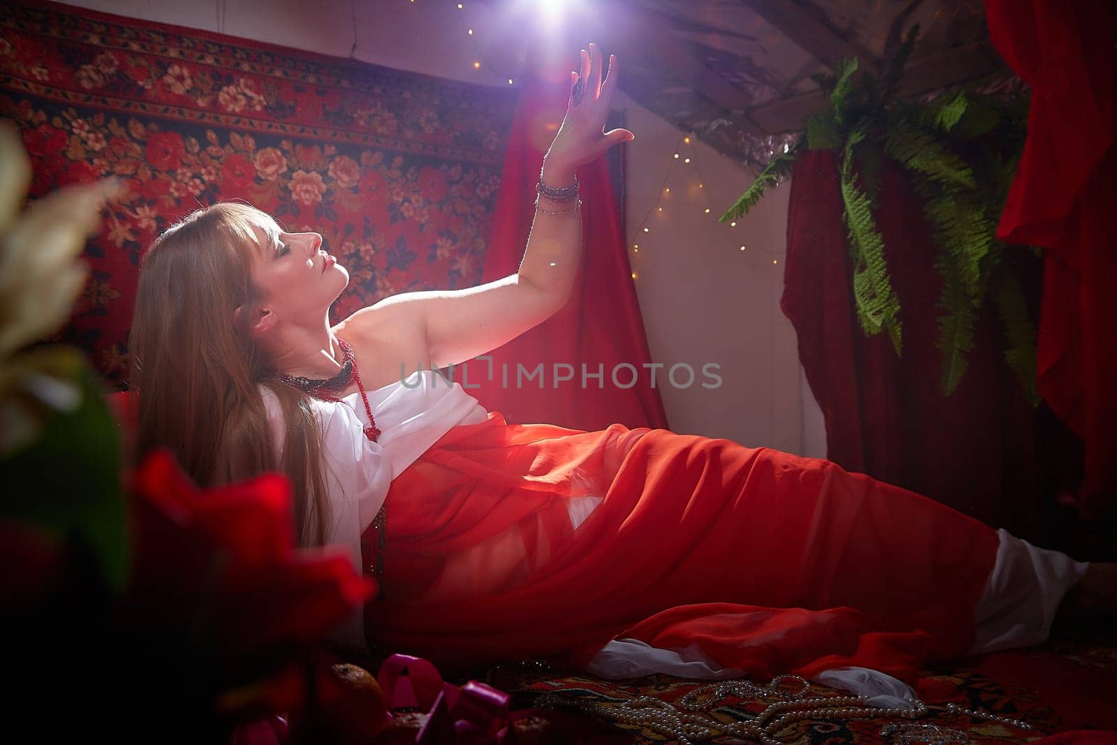 Beautiful European girl looking like Arab woman in red room with rich fabrics and carpets in sultan harem. Photo shoot of an oriental style odalisque. A model poses in sari as an indian woman in india by keleny