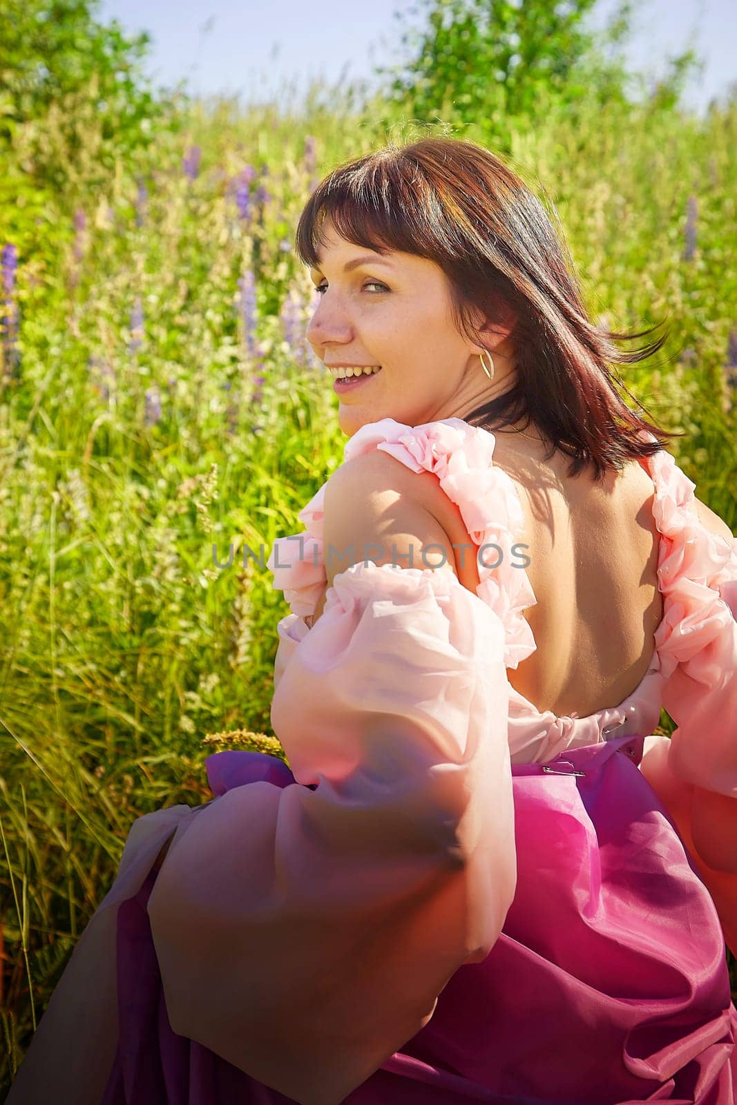 Beautiful girl in lush pink ball gown in green field during blooming of flowers and blue sky on background. Model posing on nature landscape as princess from fary tale