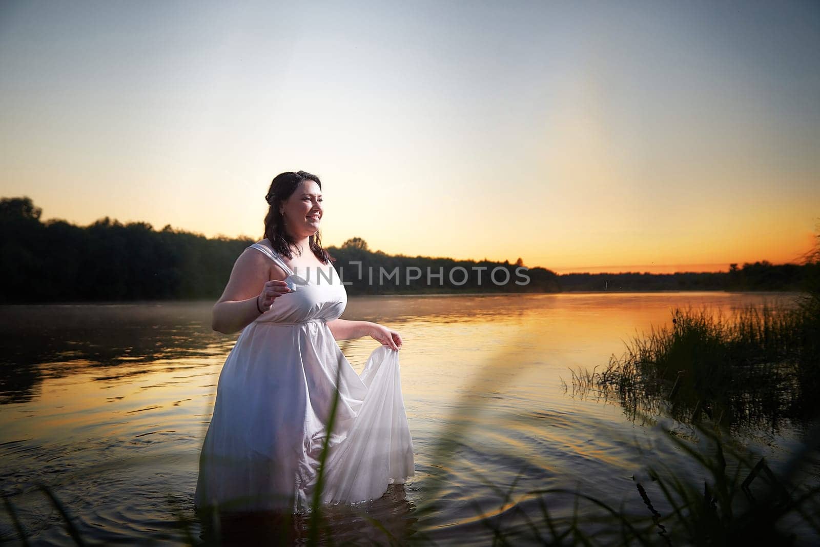 Slavic plump plump chubby girl in long white dress on the feast of Ivan Kupala with flowers and water in a river or lake on a summer evening by keleny