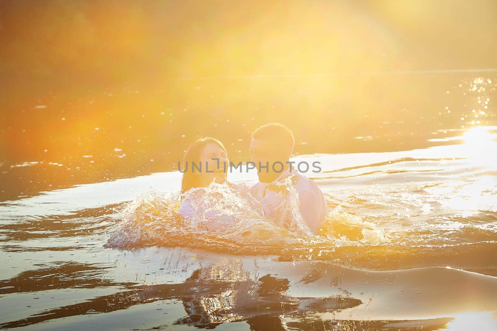 Beautiful adult couple has fun in nature in the water in a river or lake in the summer evening at sunset. A guy and a girl swim and relax outdoors in clothes in white shirts and jeans