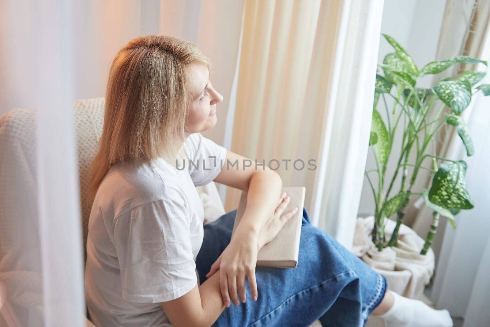 Young blonde woman with book on chair near window at home. Student girl is resting and reading textbook at home. Nice atmosphere. The concept of cozy home