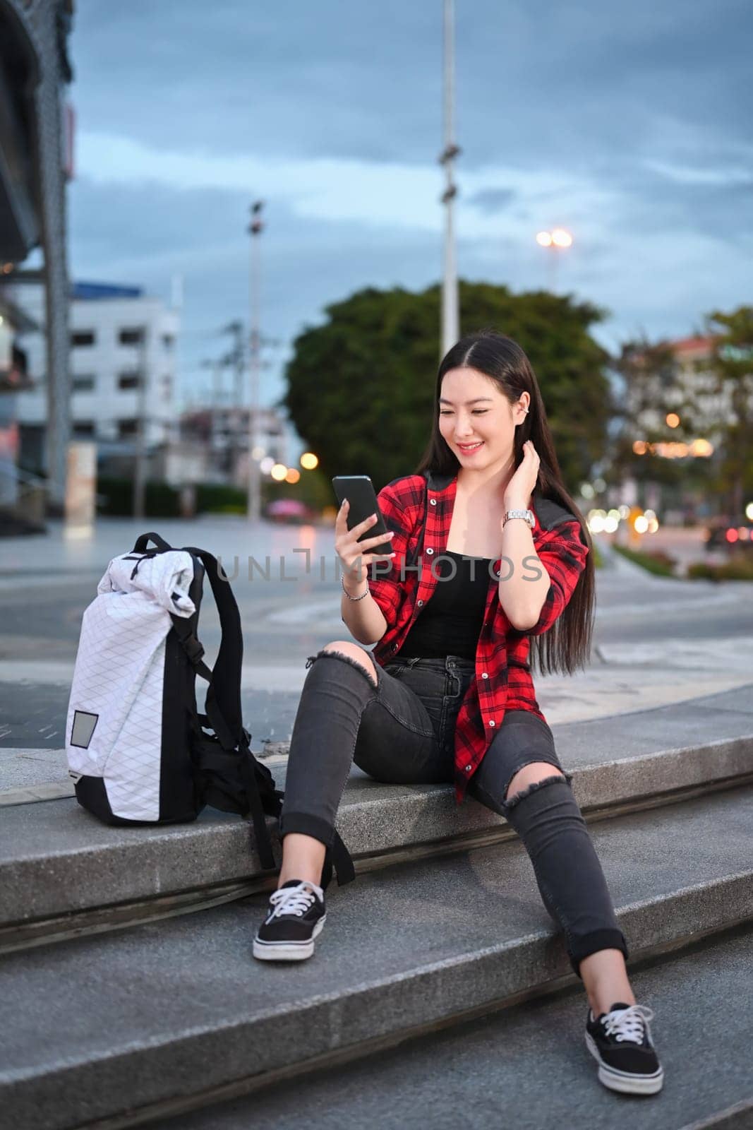 Portrait of smiling young woman sitting on stairs in the city at night and using smart phone.