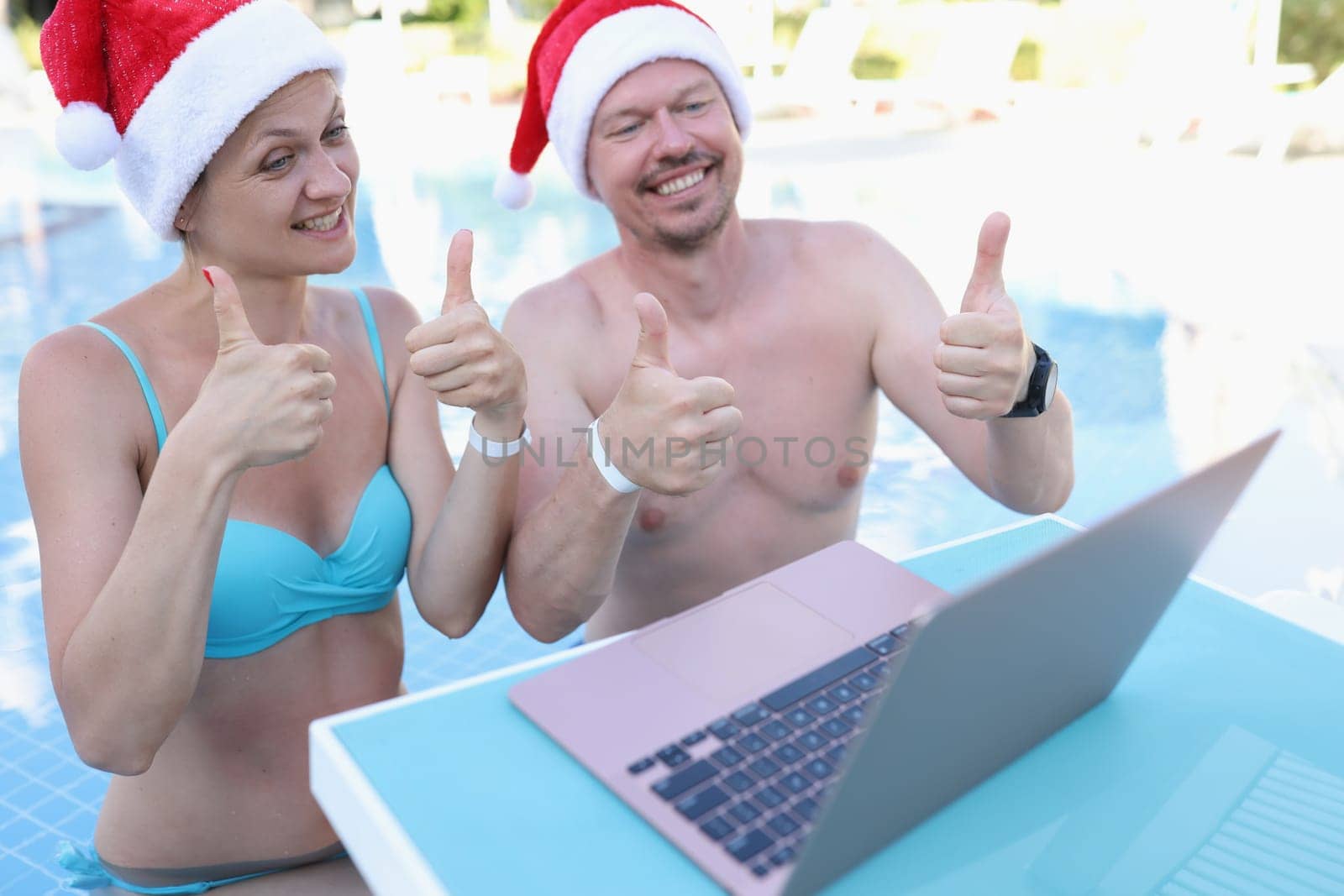 Family of bloggers in santa claus hats show thumbs up in laptop monitor standing in pool. New Year holidays tourism and travel concept