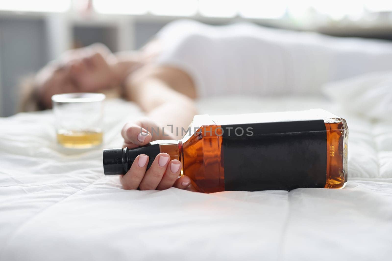 Woman lies in white bed with bottle of whiskey in hand. Alcohol addiction stress and depression concept