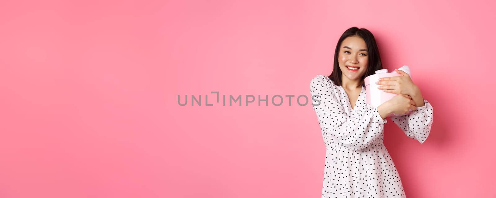 Lovely asian woman hugging her gift and smiling thankful, receive valentines day present, standing over pink background by Benzoix