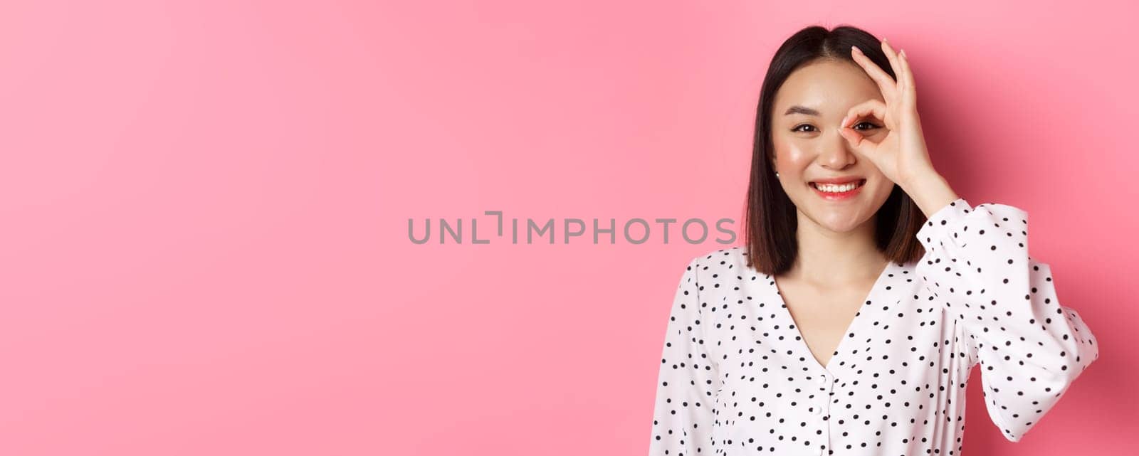 Beauty and lifestyle concept. Close-up of cute smiling asian girl showing okay sign on eye, standing over pink background in dress by Benzoix