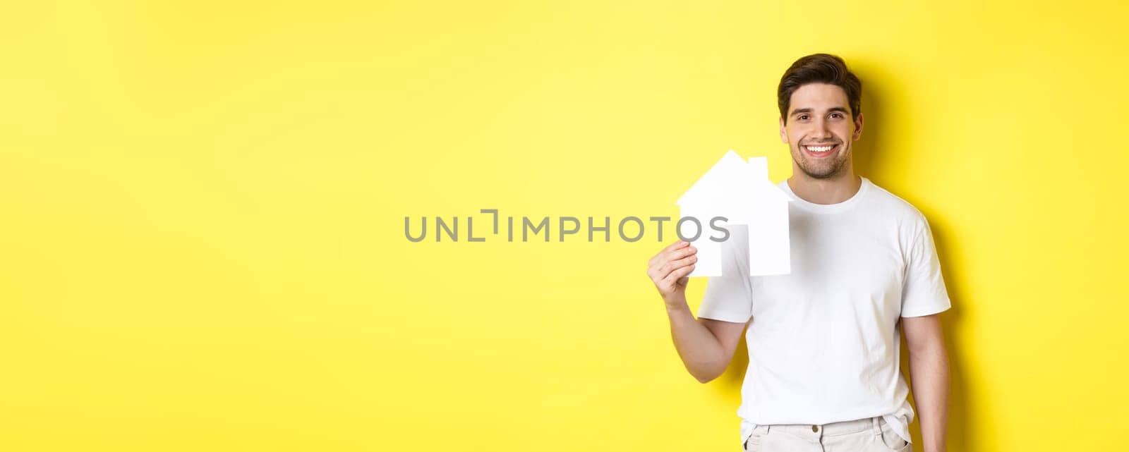 Real estate concept. Young man in white t-shirt holding paper house model and smiling, searching for apartment, standing over yellow background by Benzoix