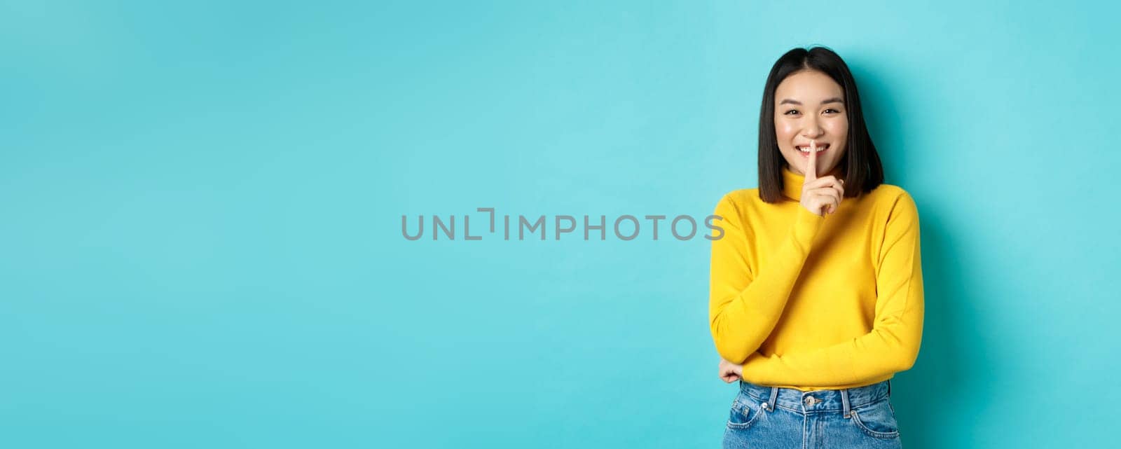 Cheerful asian girl asking to be queit, shushing at camera and smiling, keeping secret, standing against blue background by Benzoix