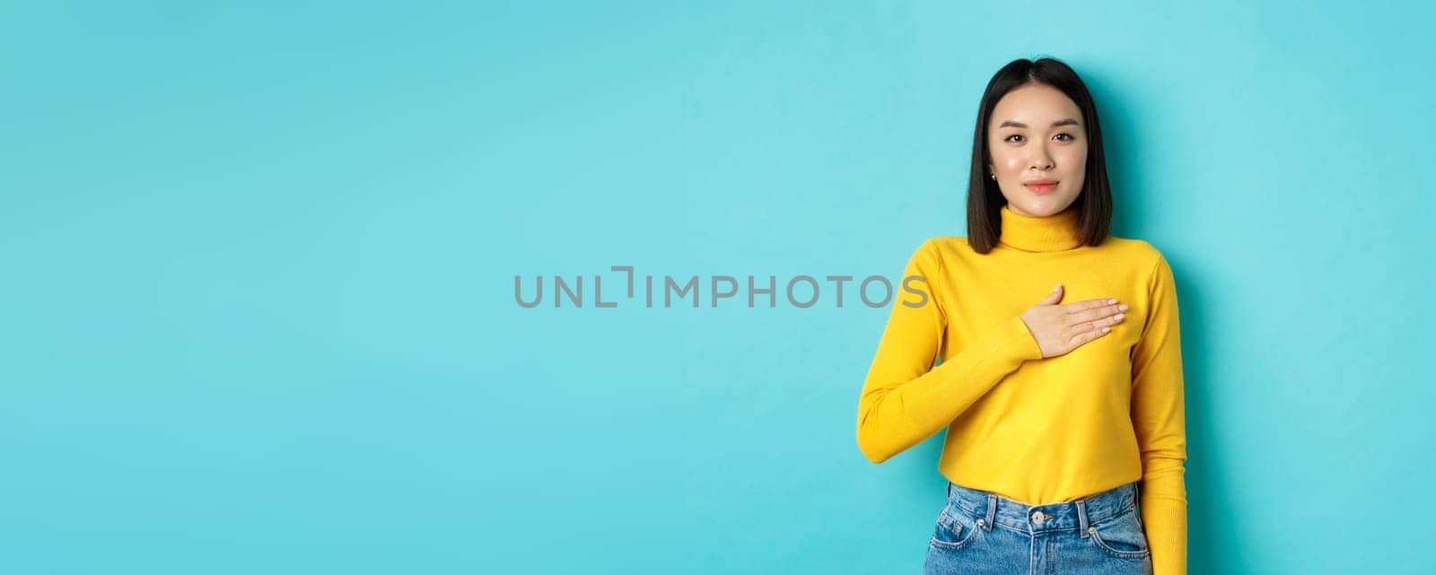 Image of proud smiling asian woman holding hand on heart, showing respect to national anthem, standing over blue background by Benzoix