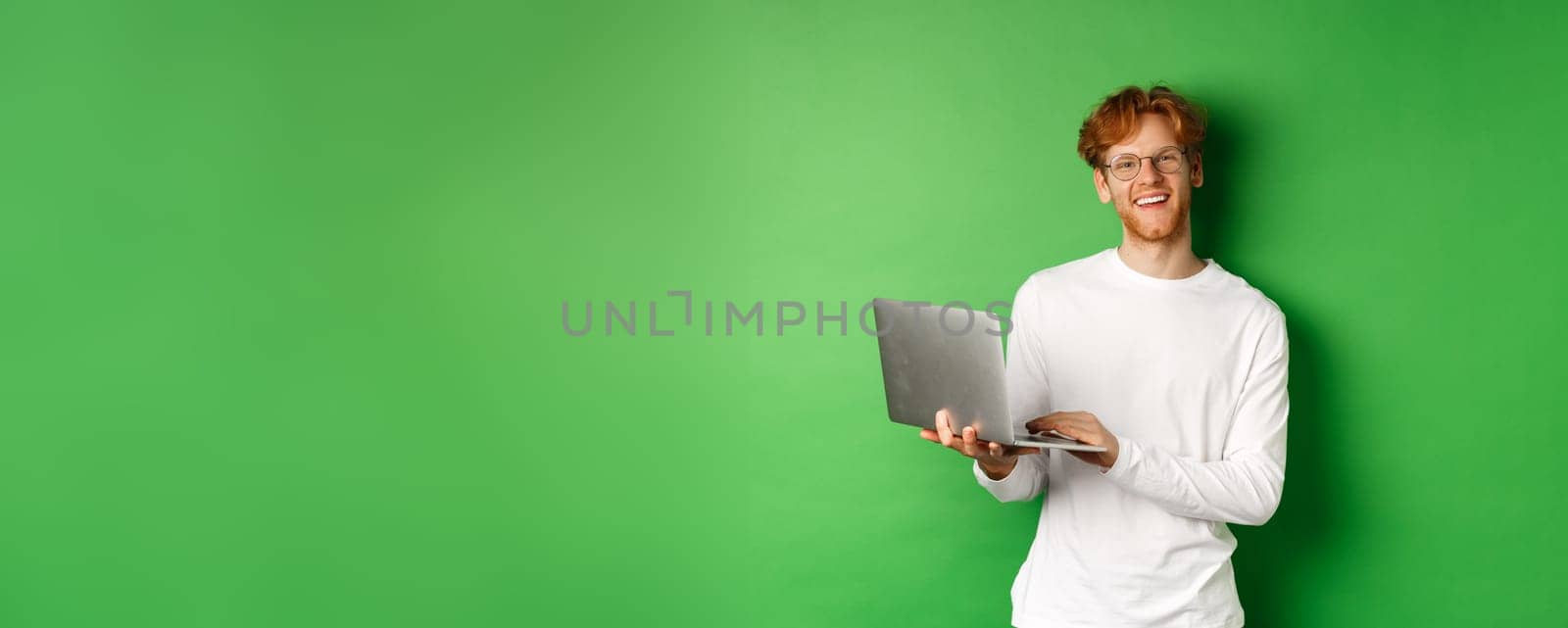 Cheerful redhead freelancer in glasses smiling at camera, working on laptop while standing over green background by Benzoix