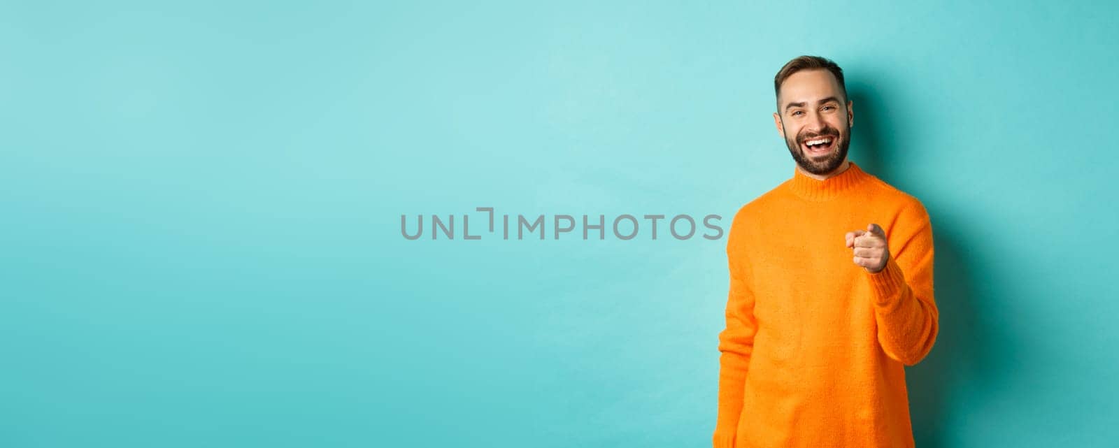 Handsome man laughing and pointing at camera, nodding in approval, agree with you, standing over light blue background.