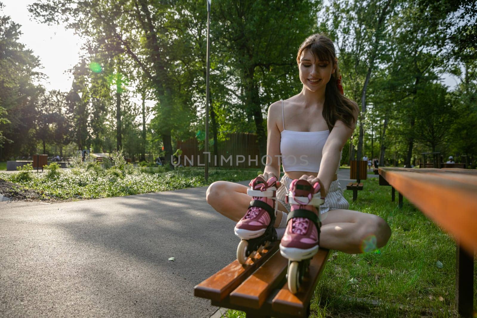 Spending time in the park on a sunny summer day. A girl is sitting on a brown bench with her legs crossed. She is holding a pair of rollerblades in front of her. Lots of trees and grass in the background.