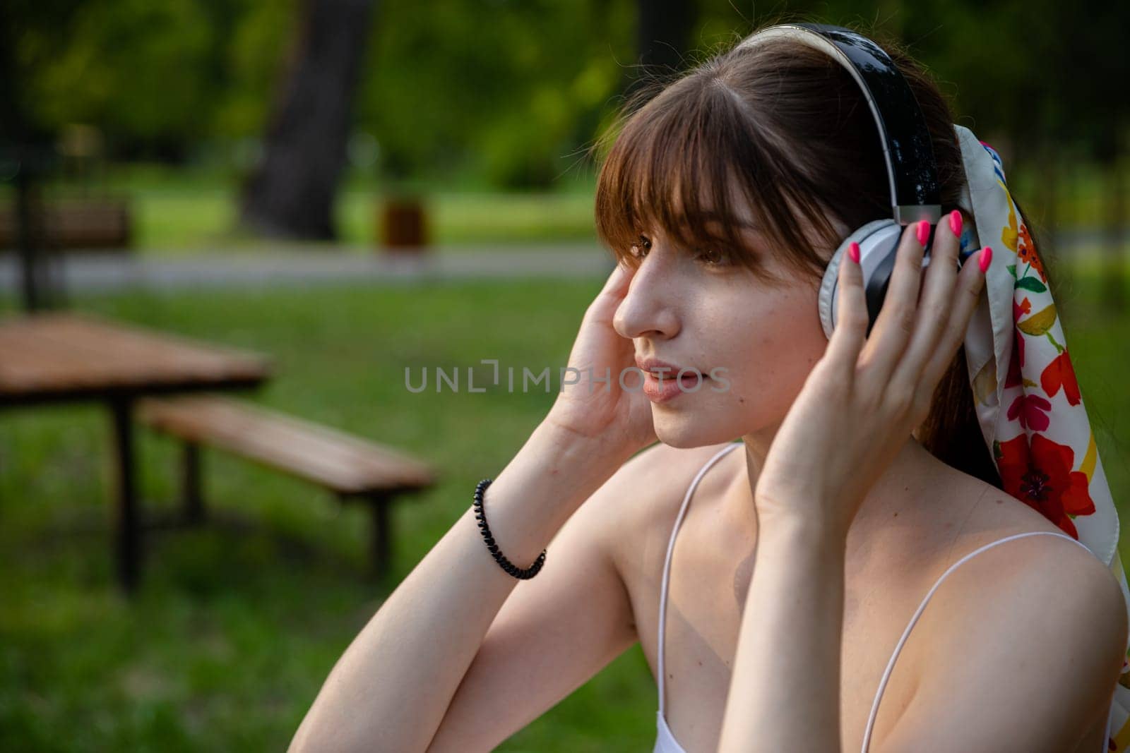 A close-up of a young girl's face. She wears wireless headphones on her head. The woman looks ahead and listens to music. She has a ponytail hairstyle with a long fringe. In the background are fuzzy lawns and brown wooden benches.