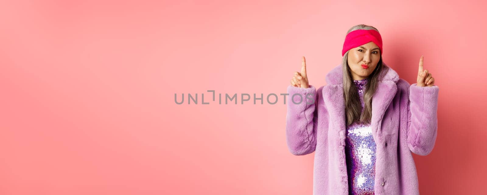 Disappointed stylish asian lady pointing fingers up. Senior woman in trendy fake fur coat grimacing skeptical and pointing fingers up, dislike promo, standing over pink background by Benzoix