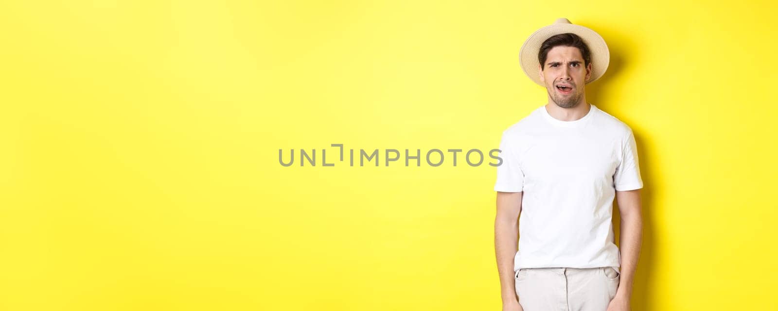 Concept of tourism and summer. Confused guy traveller in straw hat, looking puzzled, cant understand something, standing over yellow background by Benzoix