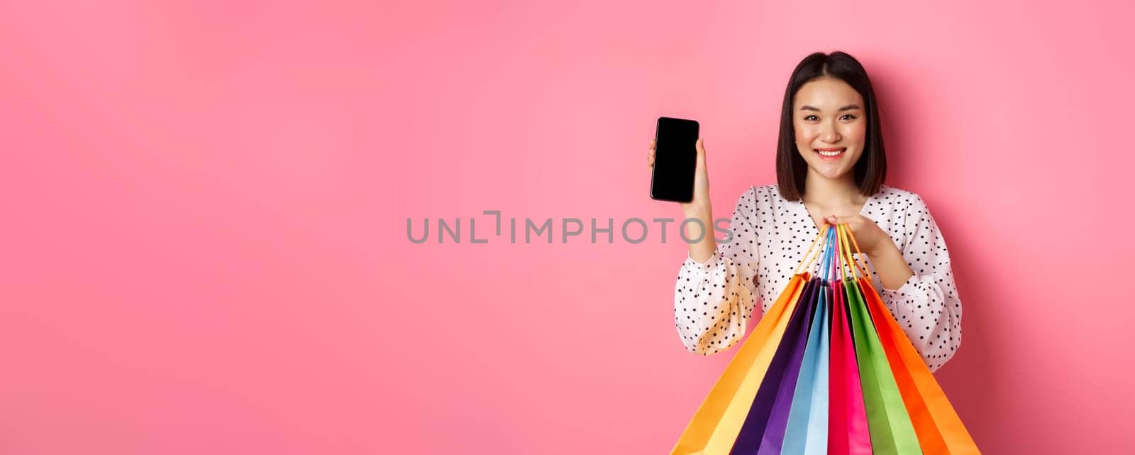 Attractive asian woman showing smartphone app and shopping bags, buying online via application, standing over pink background.