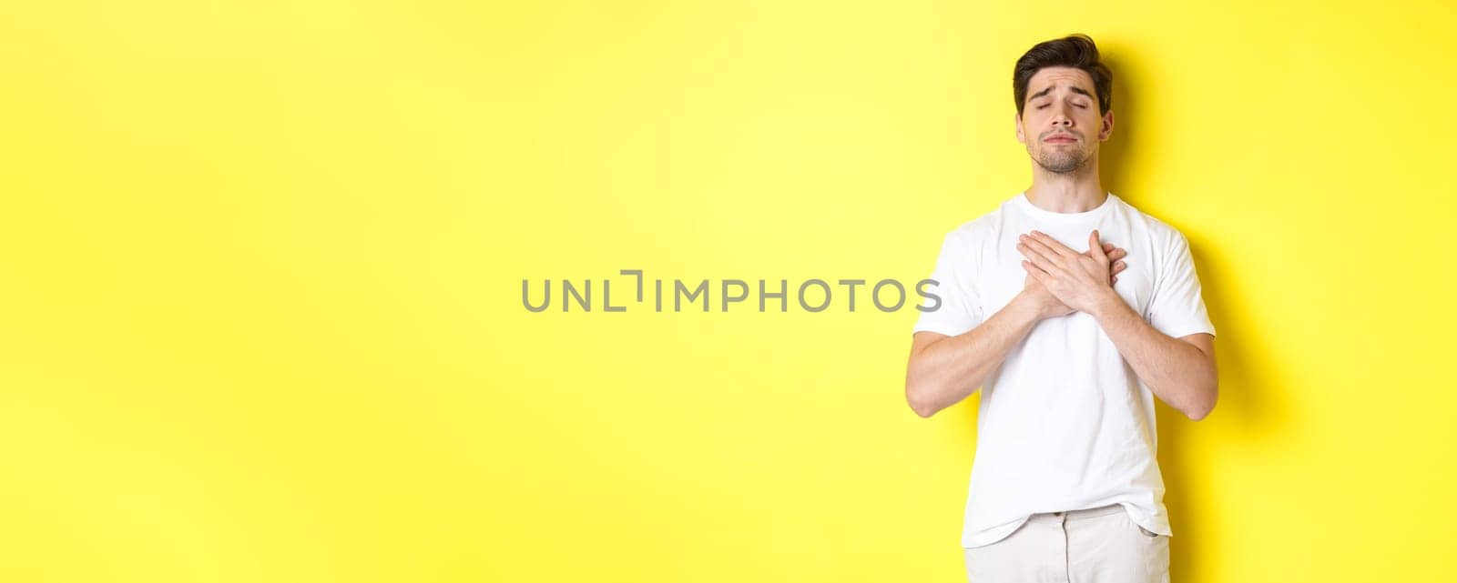 Romantic and nostalgic man holding hands on heart, close eyes and remember something, standing over yellow background.