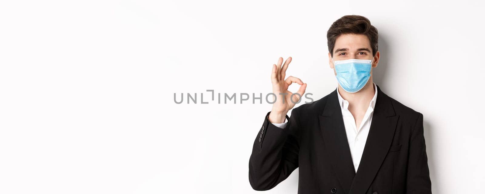 Concept of covid-19, business and social distancing. Close-up of good-looking businessman in trendy suit and medical mask, showing okay sign, recommending something, white background.
