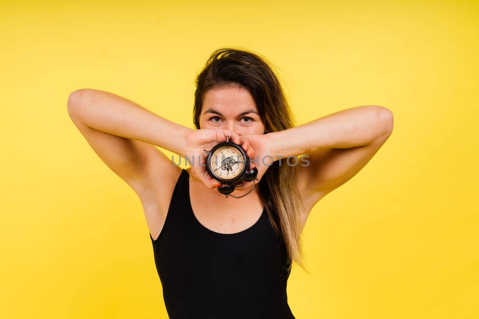 Female holding alarm clock, isolated on yellow. Young model in a black swimsuit.