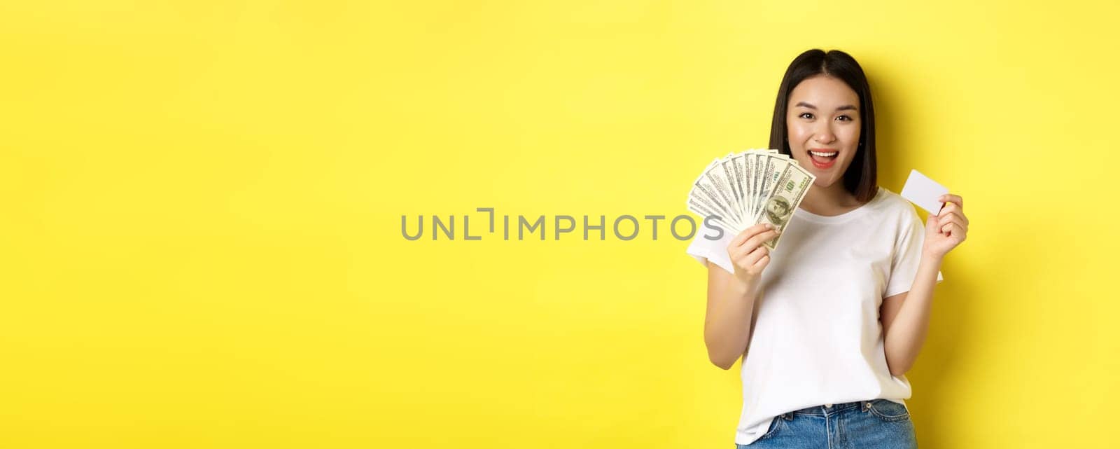 Beautiful asian woman with short dark hair, wearing white t-shirt, showing money in dollars and plastic credit card, standing over yellow background.