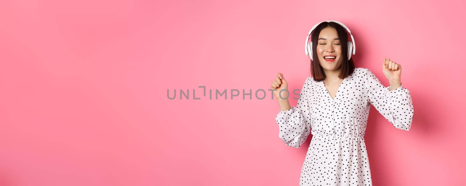 Happy young asian woman dancing and having fun, listening music in headphones, standing over pink background. Copy space