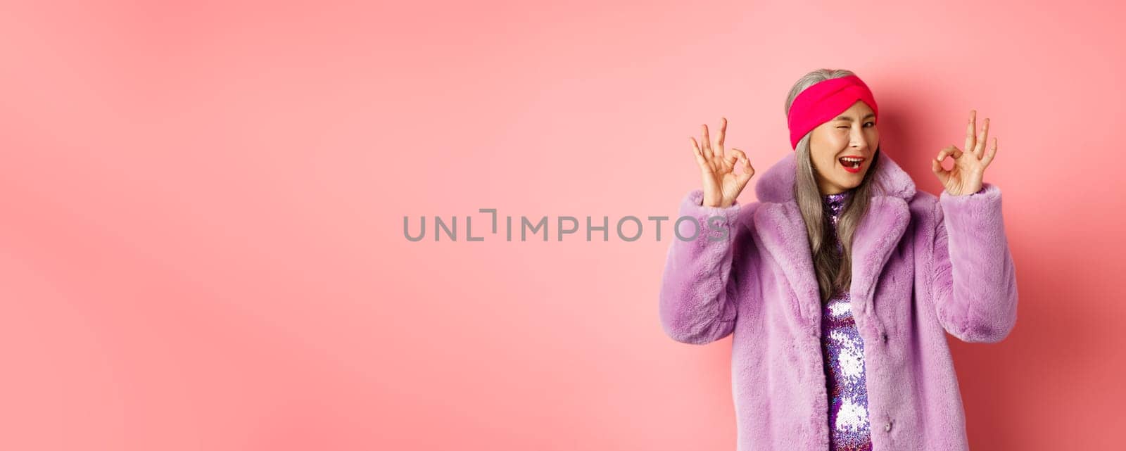 Fashion and shopping concept. Stylish asian granny saying all okay and winking. Trendy senior woman in winter fake-fur coat show OK sign, praising good product, pink background by Benzoix