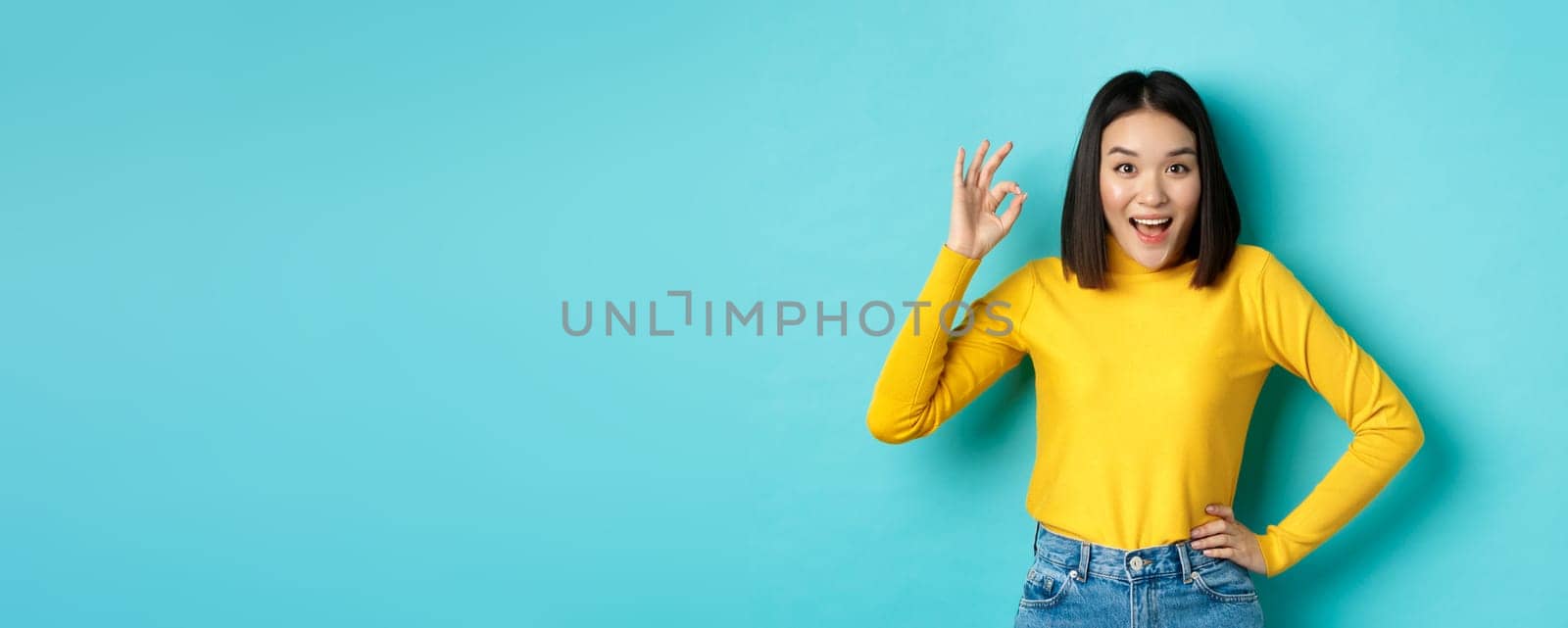 Amazed korean girl showing okay gesture and smiling at camera, approve and recommend special deal, standing over blue background by Benzoix