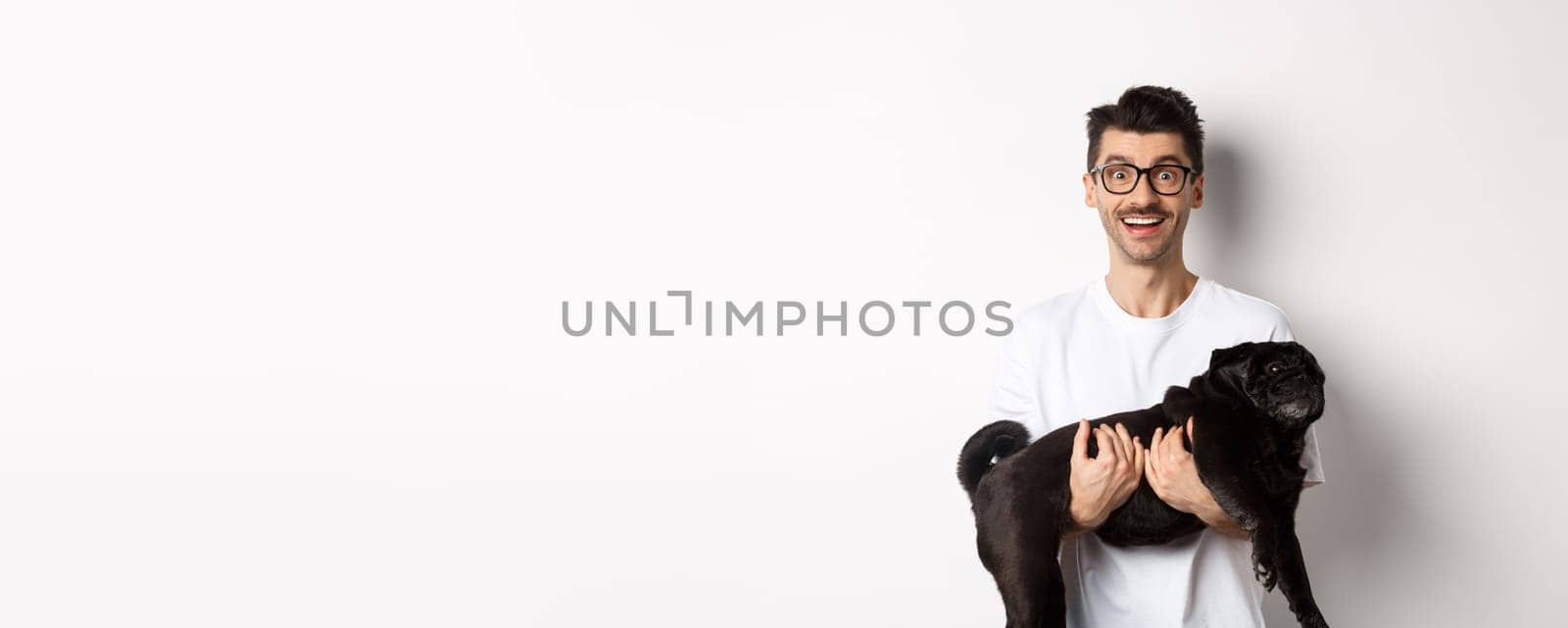Happy hipster in glasses holding cute black pug and smiling, dog owner staring at camera with amazed smile, standing over white background.