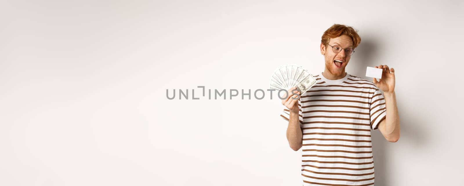 Shopping and finance concept. Young redhead man in glasses choosing plastic credit card, showing dollars and smiling, standing over white background.