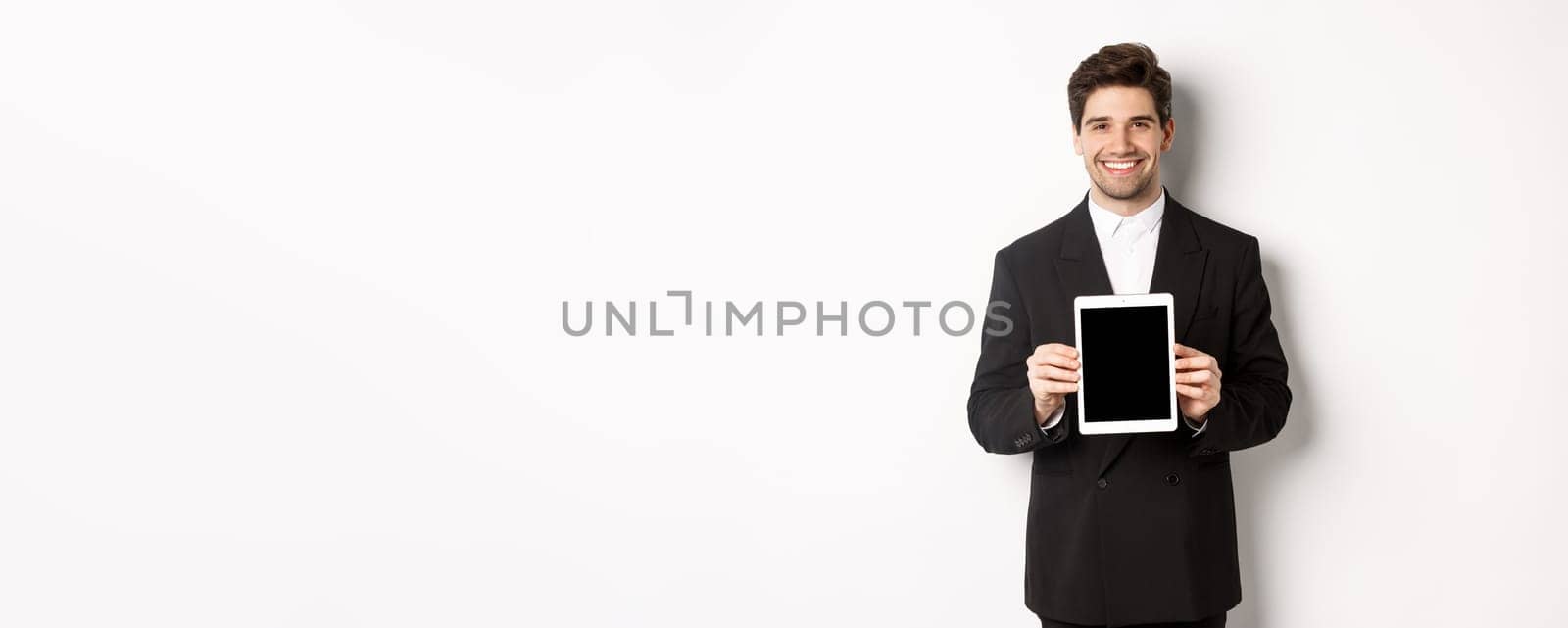 Portrait of handsome businessman in trendy suit, showing digital tablet screen and smiling, standing against white background.