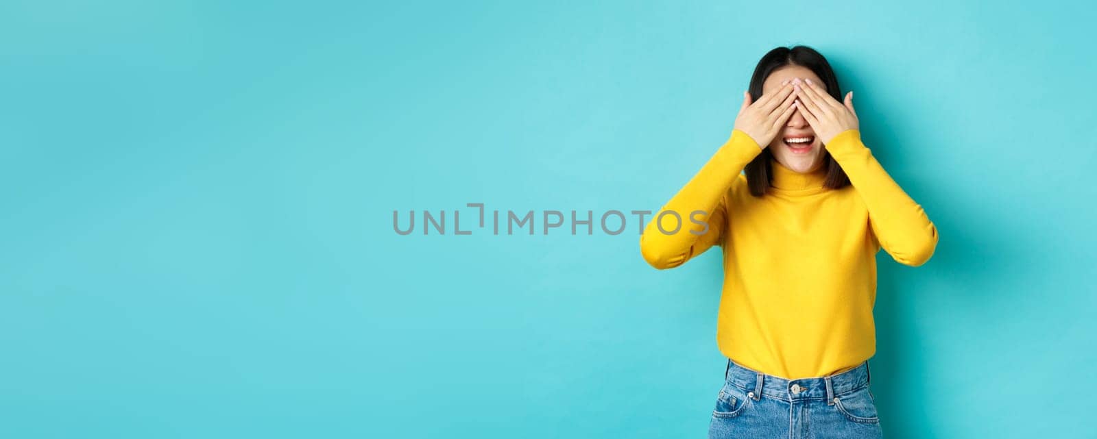 Cheerful asian girl in yellow pullover waiting for surprise, playing hide n seek and smiling, expecting gift with eyes closed, standing over blue background.