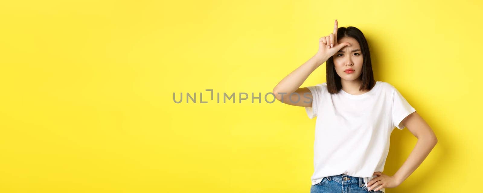 Serious and arrogant asian woman mocking lost team, showing loser sign on forehead and stare at camera confident, standing over yellow background.
