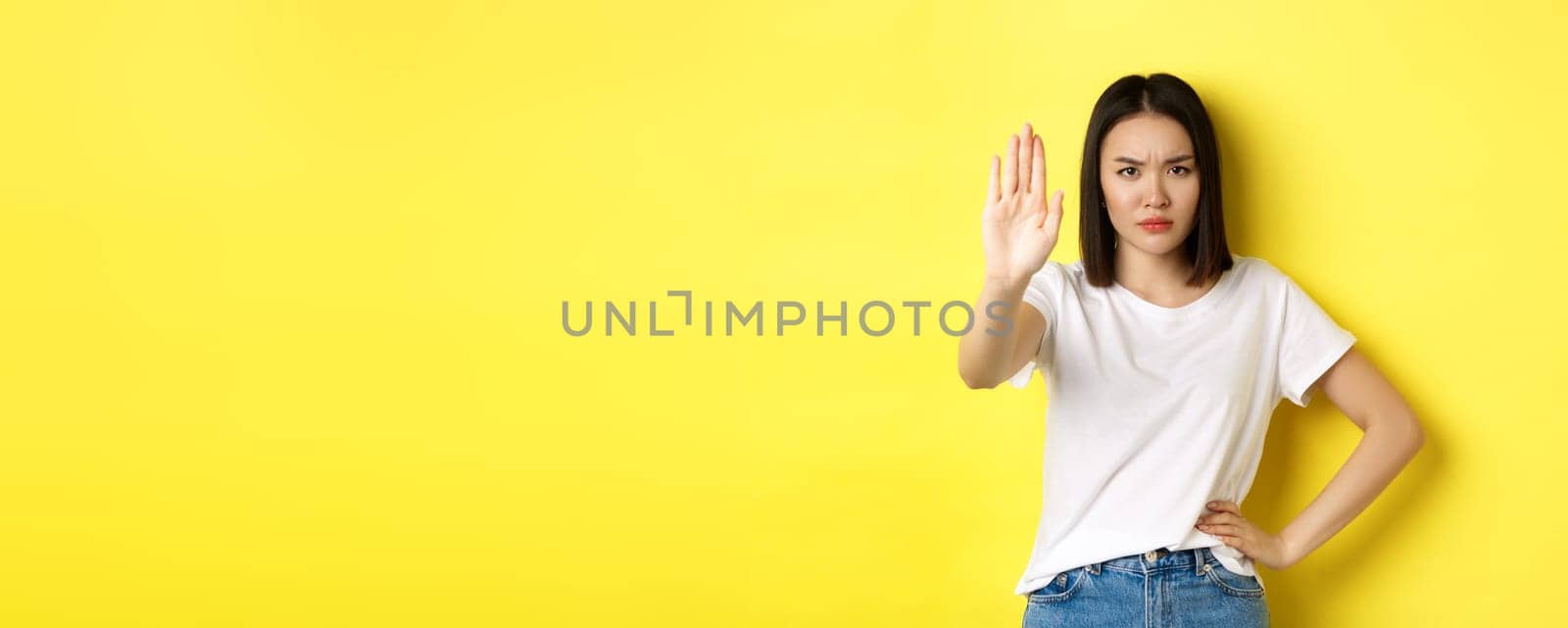 Confident and serious asian woman tell no, showing stop gesture to prohibit and warn, disagree with someone, standing upset against yellow background by Benzoix