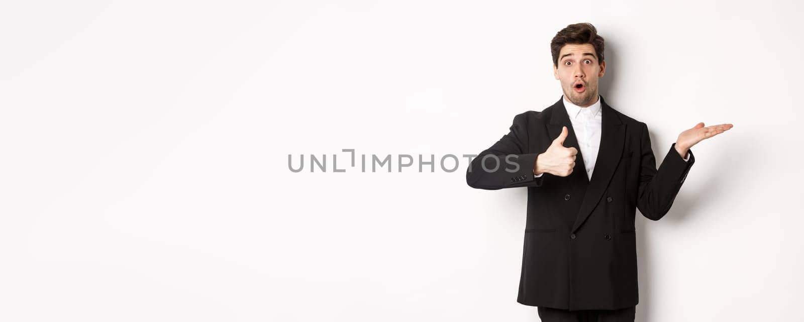 Portrait of hadnsome bearded man in formal suit, showing thumb-up and holding product in hand over white copy space, recommending product, standing over white background.