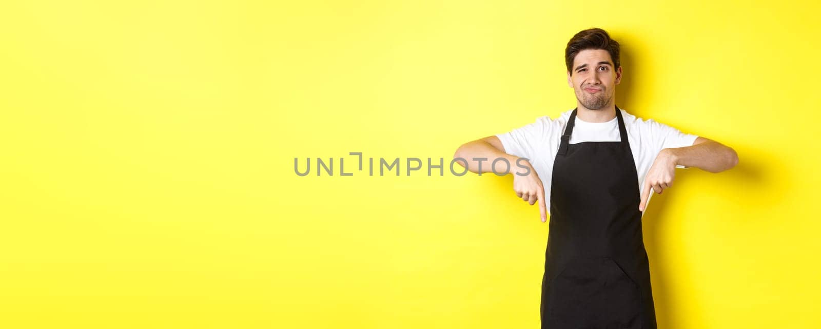 Skeptical barista in black apron pointing fingers down at bad product, looking displeased and unamused, standing over yellow background.