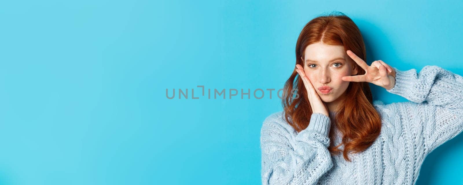 Close-up of beautiful and sassy girl with red hair, showing peace kawaii sign and gazing at camera, standing over blue background.