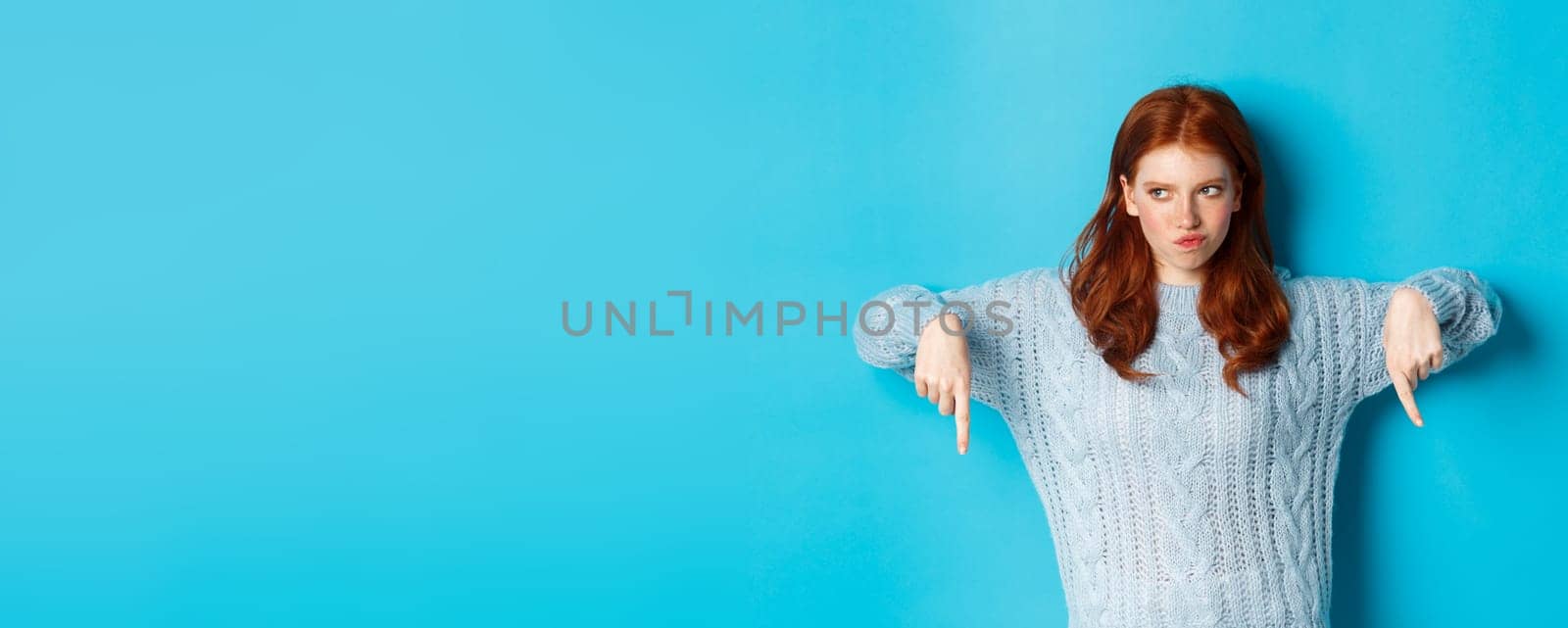 Winter holidays and people concept. Indecisive redhead girl in sweater pointing fingers down and thinking, having doubts, standing over blue background.