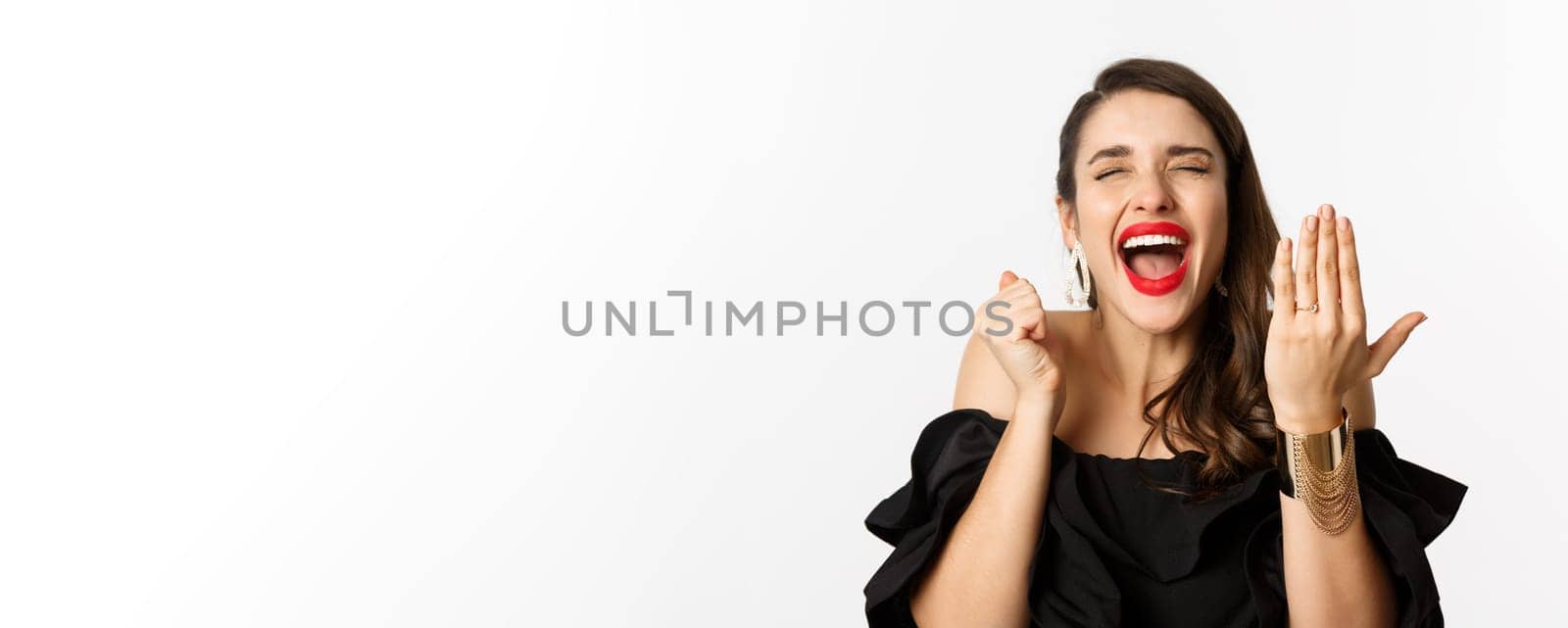 Fashion and beauty concept. Excited and happy woman said yes, showing hand with engagement ring and shouting joyful, become bride, standing over white background.