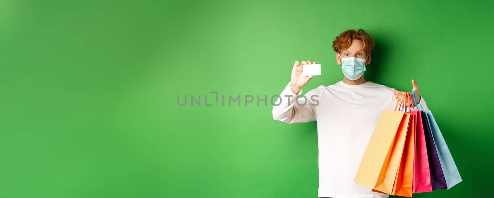 Handsome young man in medical mask, showing plastic credit card and shopping bags with items purchased with discount, green background.
